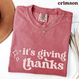 May include: Crimson-colored t-shirt with the phrase "it's giving thanks" in a white, stylized font. The shirt has a crew neck and a small tag. A silver necklace with a round pendant is draped across the shirt.