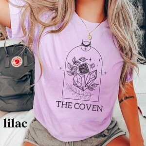May include: Lilac t-shirt with a graphic of a potion bottle, crystals, and leaves inside a dome. The text "THE COVEN" is printed below the graphic.