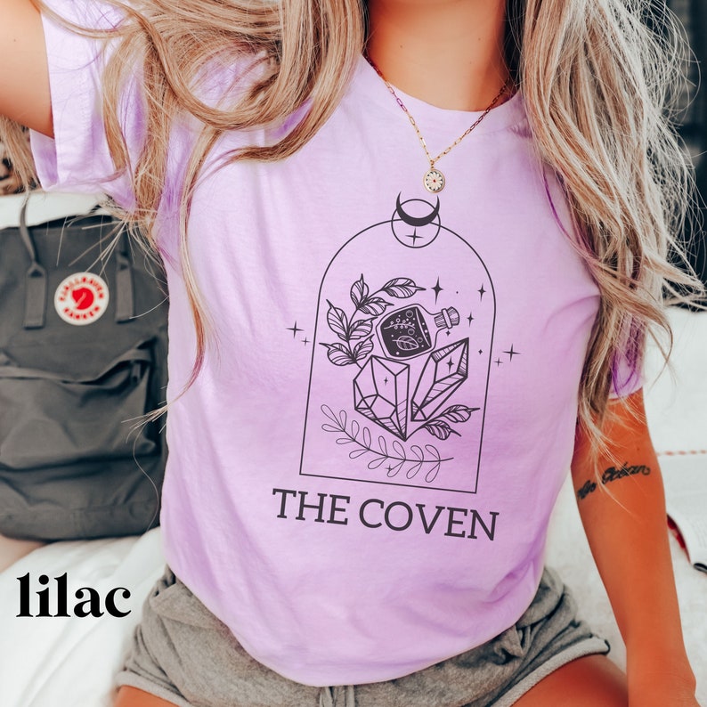 May include: Lilac t-shirt with a graphic of a potion bottle, crystals, and leaves inside a dome. The text "THE COVEN" is printed below the graphic.