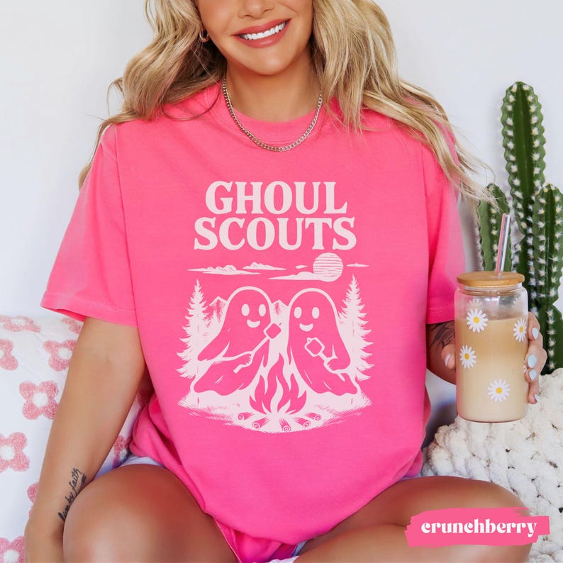 Puede incluir: Una camiseta rosa brillante con las palabras "Ghoul Scouts" en blanco, sobre un gr&aacute;fico de dos figuras de fantasmas alrededor de una fogata. El dise&ntilde;o incluye &aacute;rboles, un sol y una fogata. Una prenda de vestir informal.