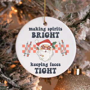 May include: A white, round Christmas ornament with the text "making spirits BRIGHT keeping faces TIGHT". The ornament features a Santa Claus graphic wearing star-shaped glasses and holding syringes. The ornament is hanging from a gold string.