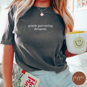 Op de afbeelding: Een donkergrijs T-shirt met de tekst "gentle parenting dropout" in wit gedrukt op de voorkant. Het shirt wordt gedragen door een persoon met lang blond haar.
