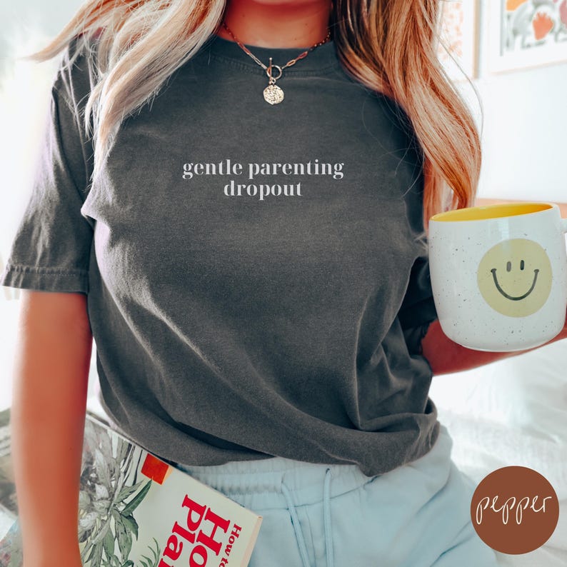 Op de afbeelding: Een donkergrijs T-shirt met de tekst "gentle parenting dropout" in wit gedrukt op de voorkant. Het shirt wordt gedragen door een persoon met lang blond haar.
