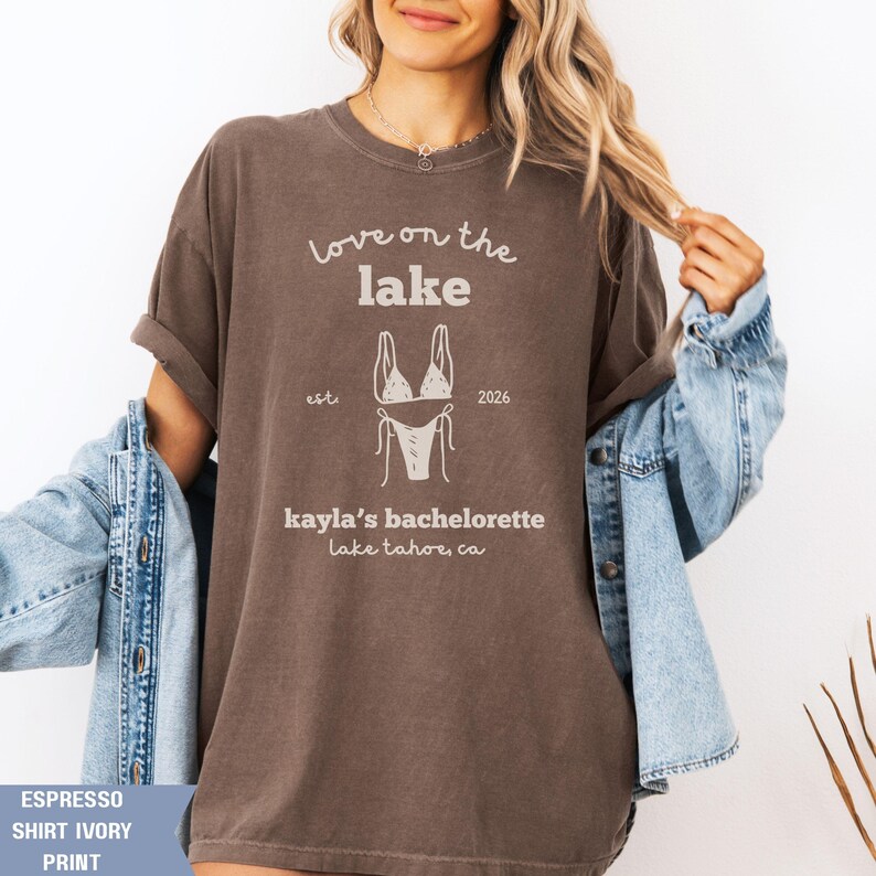 May include: Espresso-colored t-shirt with the text "Love on the Lake" and a bikini graphic. Below the graphic, it reads "kayla's bachelorette Lake Tahoe, CA." The shirt has rolled-up sleeves and is paired with a denim jacket.