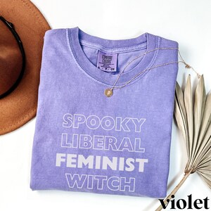 May include: A purple t-shirt with the text "Spooky Liberal Feminist Witch" in white outlined letters. The shirt is folded and laid on a white surface with a brown hat and a dried palm leaf.