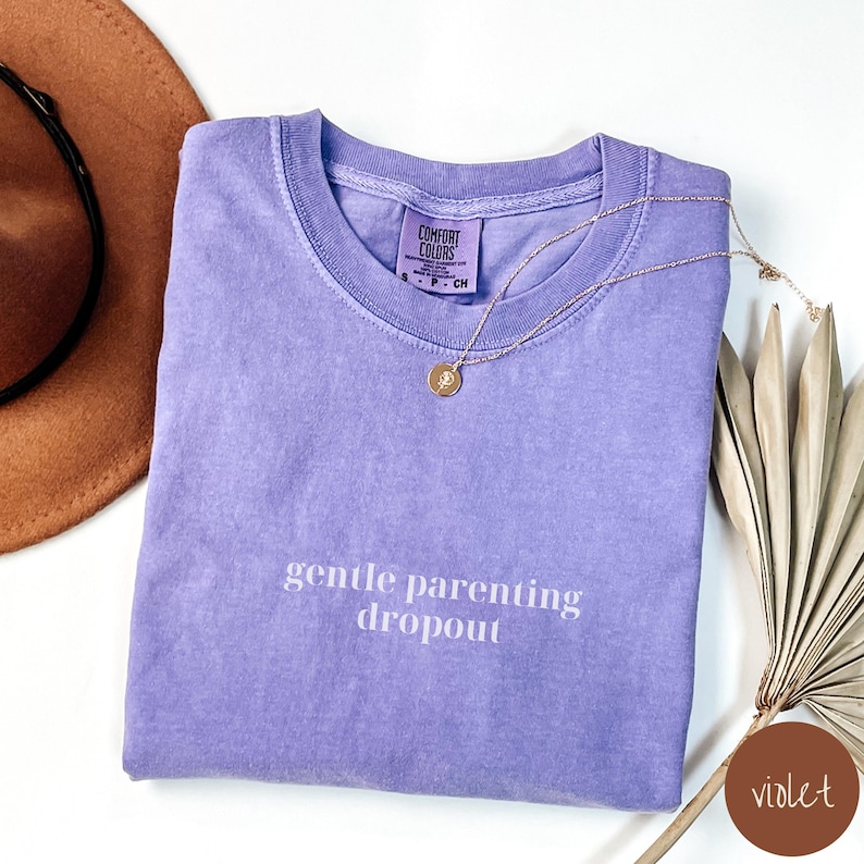 Op de afbeelding: Een paars T-shirt met de tekst "gentle parenting dropout" gedrukt op de voorkant. Het shirt is gevouwen en ligt plat op een wit oppervlak.