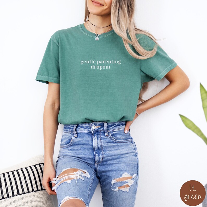 Op de afbeelding: Een vrouw draagt een lichtgroen T-shirt met de tekst "gentle parenting dropout" in wit. Ze draagt een blauwe jeans met scheuren in de knie&euml;n.