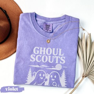Puede incluir: Una camiseta violeta Comfort Colors con la frase "GHOUL SCOUTS" en blanco. El dise&ntilde;o incluye dos figuras de fantasmas, &aacute;rboles y un sol. Tambi&eacute;n se ven un collar dorado y un sombrero marr&oacute;n.