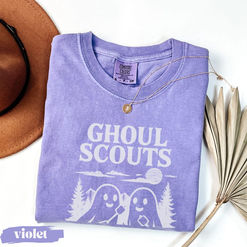 Puede incluir: Una camiseta violeta Comfort Colors con la frase "GHOUL SCOUTS" en blanco. El dise&ntilde;o incluye dos figuras de fantasmas, &aacute;rboles y un sol. Tambi&eacute;n se ven un collar dorado y un sombrero marr&oacute;n.
