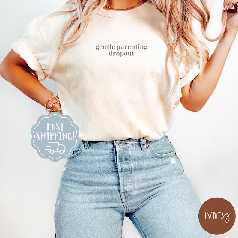 Op de afbeelding: Een wit T-shirt met de tekst "gentle parenting dropout" erop gedrukt. Het model draagt het shirt met een blauwe jeans.