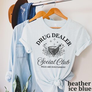 May include: A light blue t-shirt with the text "Drug Dealer Social Club Pills Are Our Passion!" in black. The text is surrounded by a black and white illustration of a mortar and pestle with flowers and stars.