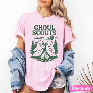 Puede incluir: Camiseta rosa claro con la frase "GHOUL SCOUTS" en verde. El dise&ntilde;o presenta dos fantasmas asando malvaviscos sobre una fogata, con &aacute;rboles y un sol. La camiseta es de estilo casual y de manga corta.
