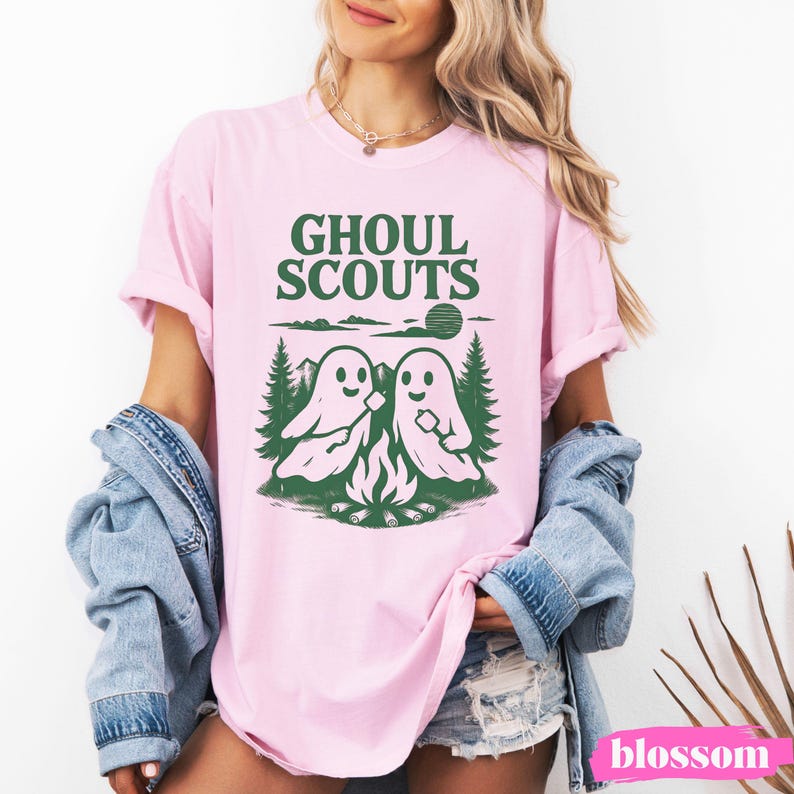 Puede incluir: Camiseta rosa claro con la frase "GHOUL SCOUTS" en verde. El dise&ntilde;o presenta dos fantasmas asando malvaviscos sobre una fogata, con &aacute;rboles y un sol. La camiseta es de estilo casual y de manga corta.