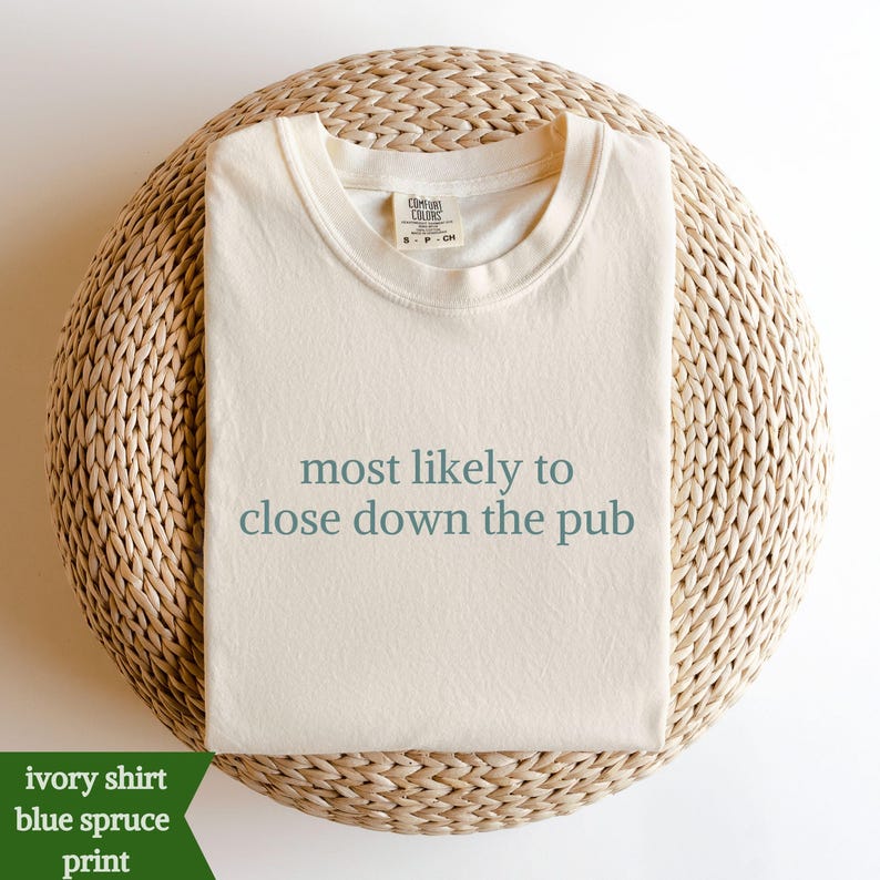 May include: An ivory-coloured t-shirt with the text "most likely to close down the pub" in blue-green lettering. The shirt is folded on a woven, circular surface. The shirt has a crew neck and short sleeves.