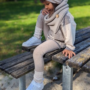 Könnte beinhalten: Ein Kind trägt eine beige Mütze mit dem Aufdruck "ciao KAKAO", einen passenden Schal und Leggings. Das Outfit umfasst ein grau-beiges Sweatshirt und weiße Turnschuhe. Das Kind sitzt auf einer Holzbank.