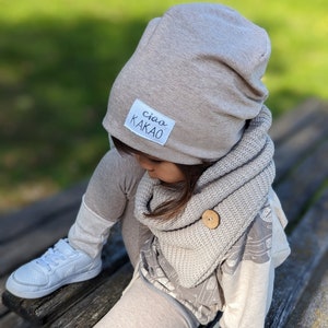 Könnte beinhalten: Ein Kind trägt eine graue Mütze mit dem Text "ciao kakao" darauf, einen grauen Strick Schal und graue Leggings. Das Kind sitzt auf einer Holzbank.