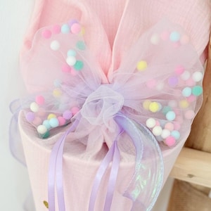 Könnte beinhalten: Eine rosa kegelförmige Geschenktüte mit einer Tüllschleife, die mit bunten Pompons verziert ist. Die Tasche ist mit lavendelfarbenen Bändern und einer transparenten, irisierenden Bordüre verziert. Der Text "wish" ist auf der Tasche zu sehen.