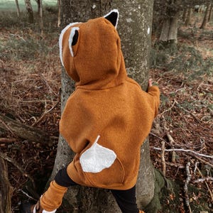 Könnte beinhalten: Ein Kind trägt einen braunen Kapuzenpullover mit einem weißen Fuchsschwanz und -ohren. Der Pullover ist aus einem weichen, flauschigen Material gefertigt. Das Kind steht in einer Waldumgebung.