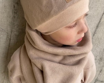 May include: A beige beanie and a matching beige neck warmer. The beanie features a square brown patch with the text "hej" in black. The neck warmer also includes a matching square brown patch with the text "hej" in black, positioned at the bottom.