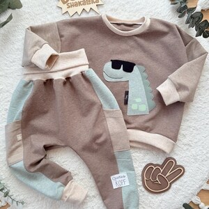 Könnte beinhalten: Ein zweiteiliges Kinder-Outfit in Braun, Beige und Hellblau. Das langärmlige Oberteil hat eine Dinosaurier-Grafik mit Sonnenbrille. Die Hose hat ein Patchwork-Design. Das Outfit enthält den Text "BOOM! Shakalka" und "Quetsch KOPF".