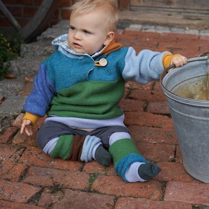 Könnte beinhalten: Ein kleines Kind trägt einen bunten, mehrfarbigen gestreiften Pullover und passende Beinstulpen und sitzt auf einer gepflasterten Terrasse. Das Kind hält einen Metalleimer.