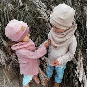 Könnte beinhalten: Zwei Kinder tragen passende rosa und beige Strickmützen und Schals. Die Mützen haben ein kleines braunes Etikett mit dem Text "Ciao Kakao" darauf. Die Kinder tragen blaue Jeans und stehen vor einem Hintergrund aus hohem Gras.