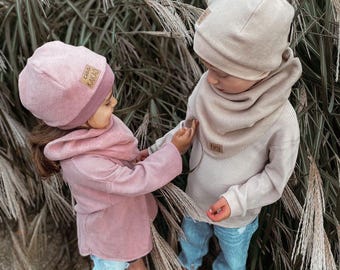 May include: Two children wearing matching pink and beige knitted hats and scarves. The hats have a small brown label with the text "Ciao Kakao" on them. The children are wearing blue jeans and are standing in front of a background of tall grass.