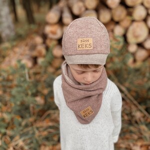 Könnte beinhalten: Ein Kind trägt ein passendes Set aus mauvefarbener Mütze und Schal. Beide Teile haben ein kleines rechteckiges Etikett mit dem Text "süß Keks". Das Kind trägt einen weißen Pullover. Im Hintergrund Holzstämme.