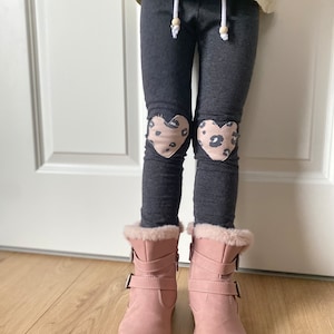 Könnte beinhalten: Graue Leggings mit einem herzförmigen Patch auf jedem Knie. Die Patches sind rosa und haben einen Leoparden-Druck. Die Leggings werden mit rosa Stiefeln getragen.
