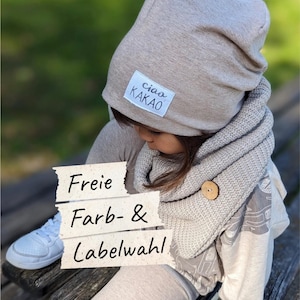 Könnte beinhalten: Eine beigefarbene Mütze mit einem weißen rechteckigen Etikett mit der Aufschrift ciao KAKAO und ein passender gestrickter Schlauchschal mit einem einzelnen Holzknopf. Ein Papieraufleger zeigt den Text Freie Farb- & Labelwahl.
