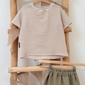 Könnte beinhalten: Ein beiges kurzärmeliges T-Shirt und eine olivgrüne Shorts, hängen an einer Holzleiter. Das T-Shirt hat einen Rundhalsausschnitt und einen strukturierten Stoff. Die Shorts haben einen elastischen Bund. Im Hintergrund ist Wimpelkette aus Jute.