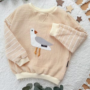 Könnte beinhalten: Hellpfirsichfarbenes Sweatshirt mit einem Möwen-Applikation. Die Möwe hat einen weißen Körper, einen grauen Flügel und einen orangefarbenen Schnabel und Beine. Die Ärmel haben beige und weiße Streifen. Der Halsausschnitt und die Manschetten sind cremefarben. Das Wort "hej" steht auf der Möwe.