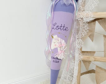 May include: A lavender fabric cone featuring the name Lotte embroidered in dark thread, a unicorn applique with yarn mane, and the text Schultüte 2026. The top is decorated with a sheer white tulle bow, small colorful pom-poms, and iridescent ribbons.