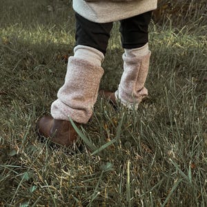 Könnte beinhalten: Ein Kind trägt braune Lederstiefel mit flauschigen rosa Beinstulpen. Das Kind trägt eine schwarze Hose und einen grau-braunen Pullover. Das Kind steht auf einer grünen Wiese.