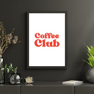 May include: A framed print with the words "Coffee Club" in red, bold, retro-style lettering on a white background. The frame is black, and the print is displayed on a dark gray surface with decorative vases and a watch.