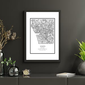 May include: A framed black and white map print of Porto, Portugal. The map shows the city's layout with streets and waterways. The print includes the city's name, coordinates, and population. The frame is black, and the print is displayed on a dark gray surface.
