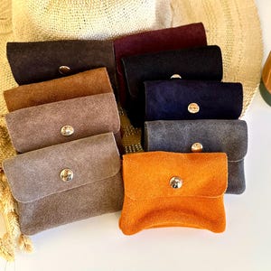 Könnte beinhalten: Eine Auswahl an kleinen Wildleder-Pouches in verschiedenen Farben, darunter Braun, Schwarz, Grau und Orange. Jede Tasche hat einen Klappenverschluss mit einem silbernen Knopf. Ein Strohhut und ein grüner Reisebecher sind im Hintergrund zu sehen.