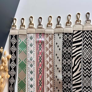 May include: A collection of patterned bag straps with gold-colored clasps. The straps feature various designs, including geometric patterns, a zebra print, and a repeating diamond pattern. The straps are in a variety of colors, including black, white, green, pink, and brown.