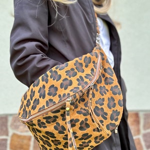 Può includere: Marsupio con stampa leopardata marrone e nera con chiusura a zip e lunga zip pull. La borsa è realizzata in materiale simile alla pelle scamosciata e ha una forma curva. La borsa è indossata sulla spalla.