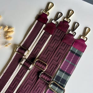 Könnte beinhalten: Vier verstellbare Taschengurte mit goldfarbenen Metallkarabinerhaken und Schiebern. Die Gurte weisen verschiedene Muster auf, darunter ein burgunderrotes Mäander-Design mit weißem Rand, strukturiertes burgunderrotes Leder und ein burgunder-grünes Karomuster.