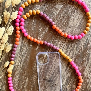 Könnte beinhalten: Ein mehrfarbiger Perlen-Handy-Anhänger mit rosa, orangefarbenen und braunen Perlen. Der Anhänger ist an einem durchsichtigen Handy-Case mit goldenen Ringen befestigt.