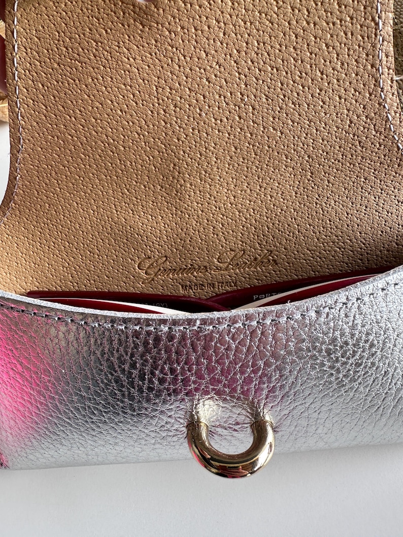 May include: Close-up of a silver leather handbag with a gold-colored clasp. The interior is a light brown color with the words "Genuine Leather Made in Italy" embossed on the leather.