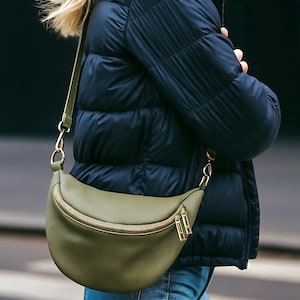 May include: An olive green crescent-shaped crossbody bag with a textured surface, featuring a gold-toned metal zipper closure and matching gold hardware. The bag has an adjustable strap attached to gold rings on each side, worn across the torso.