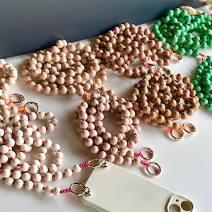 Puede incluir: Una colección de correas para teléfono con cuentas en varios colores, incluyendo blanco, beige y verde. Cada correa presenta cuentas de madera ensartadas en un cordón, con cierres metálicos dorados y detalles rosas. Las correas están diseñadas para sujetarse a una funda de teléfono.