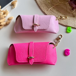 May include: Two leather eyeglass cases in pink and hot pink. Each case has a flap closure with a gold-tone ring detail and a matching gold-tone chain with a clasp. The cases are rectangular and have a textured surface.