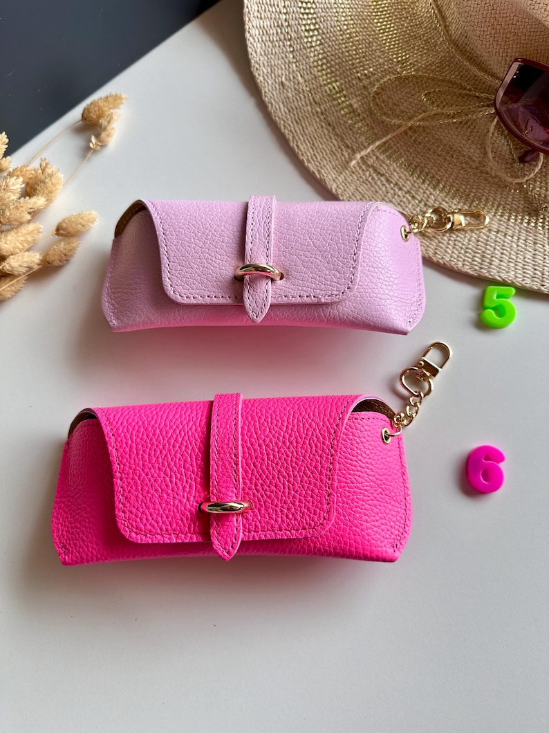 May include: Two leather eyeglass cases in pink and hot pink. Each case has a flap closure with a gold-tone ring detail and a matching gold-tone chain with a clasp. The cases are rectangular and have a textured surface.