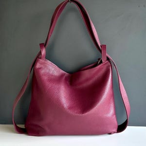 Könnte beinhalten: Eine tief magentafarbene Hobo-Tasche aus strukturiertem Leder. Die Tasche hat eine lockere, unstrukturierte Silhouette mit einem einzelnen breiten Schulterriemen und einem Reißverschluss oben. Die Oberfläche weist eine sichtbare Narbung auf.