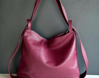 May include: A deep magenta hobo bag crafted from textured leather. The bag features a slouchy, unstructured silhouette with a single wide shoulder strap and a zippered top closure. The surface has a visible pebbled grain texture throughout.