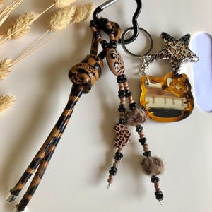 Peut inclure: Un porte-clés avec un mousqueton noir, orné d'un motif léopard. Le porte-clés comprend un charm Hello Kitty, un charm étoile, des perles et des accents de fourrure. La palette de couleurs comprend le marron, le noir et le beige.