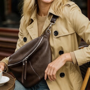 May include: A dark brown leather crossbody bag with a silver zipper and a matching strap. The bag is worn over the shoulder. The person is wearing a tan trench coat and blue jeans.
